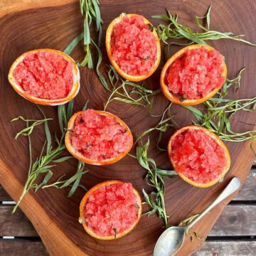 Overhead view of blood orange and tarragon granita served in halved orange shells. Six on a wooden board, surrounded by tarragon sprigs.