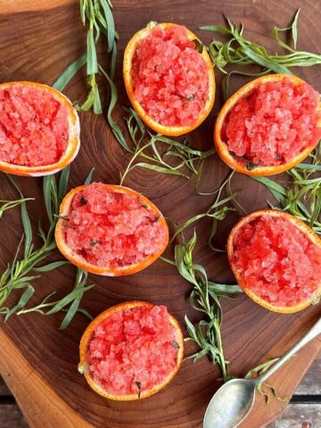 Overhead view of blood orange and tarragon granita served in halved orange shells. Six on a wooden board, surrounded by tarragon sprigs.