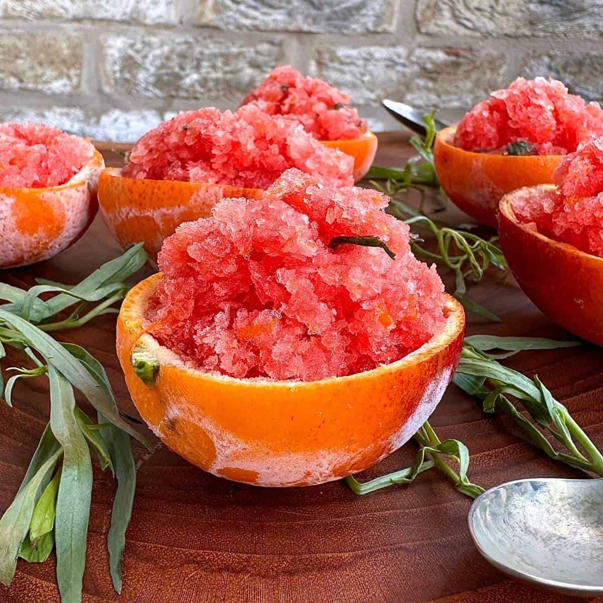 blood orange granita in hollowed out orange skins on a wooden board surrounded by sprigs of tarragon.
