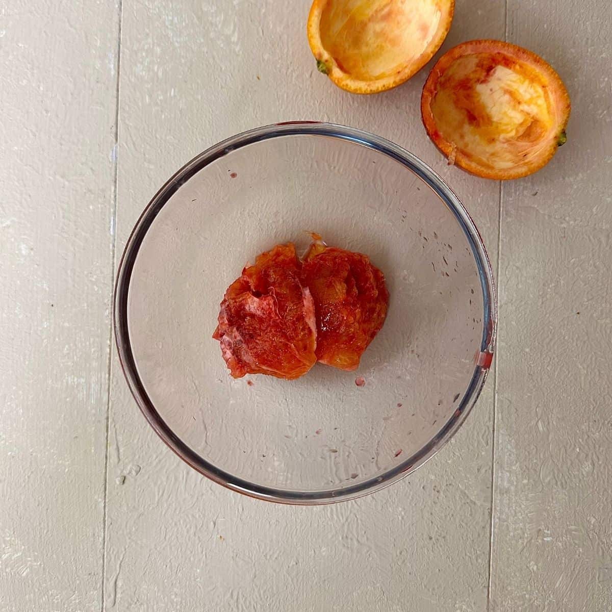 A round glass bowl containing blood orange flesh for blood orange granita recipe. Two halves of orange skin sit next to the bowl.