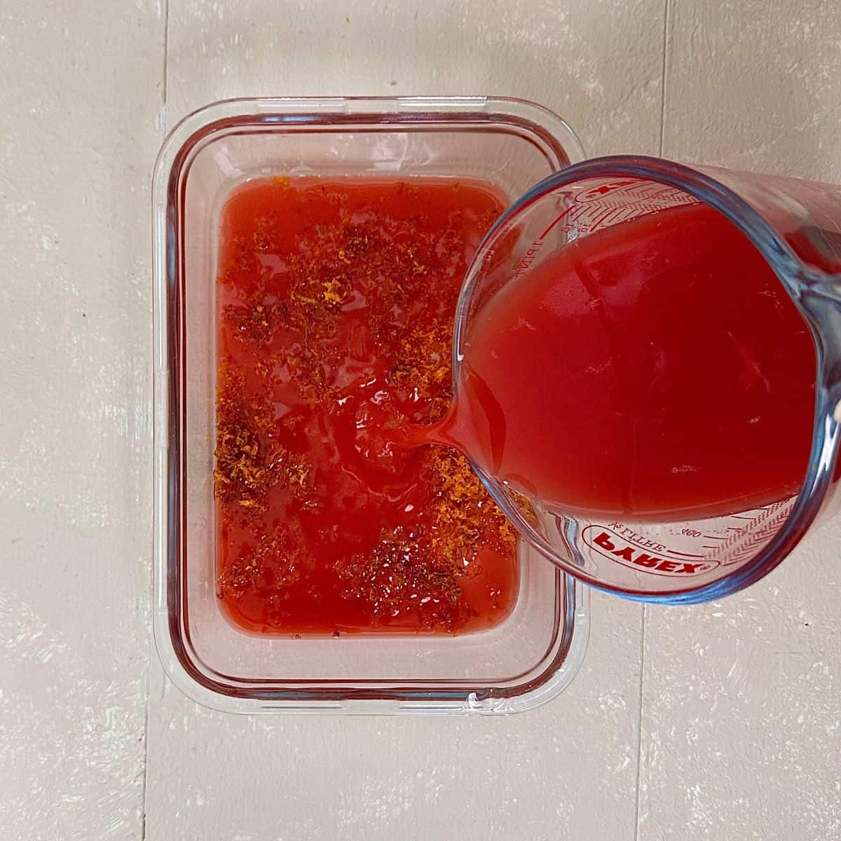 Fresh blood orange juice being poured from a glass measuring jug int a shallow glass dish containing sugar and blood orange zest for blood orange granita recipe.