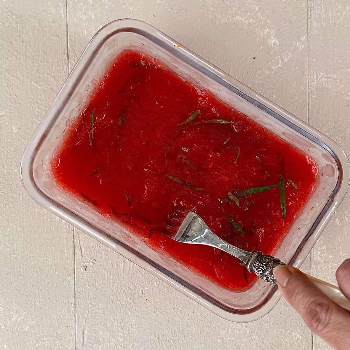 A shallow glass dish containing blood orange granita after 90 minutes freezing. A folk is being used to scrape the surface to form ice crystals before refreezing.