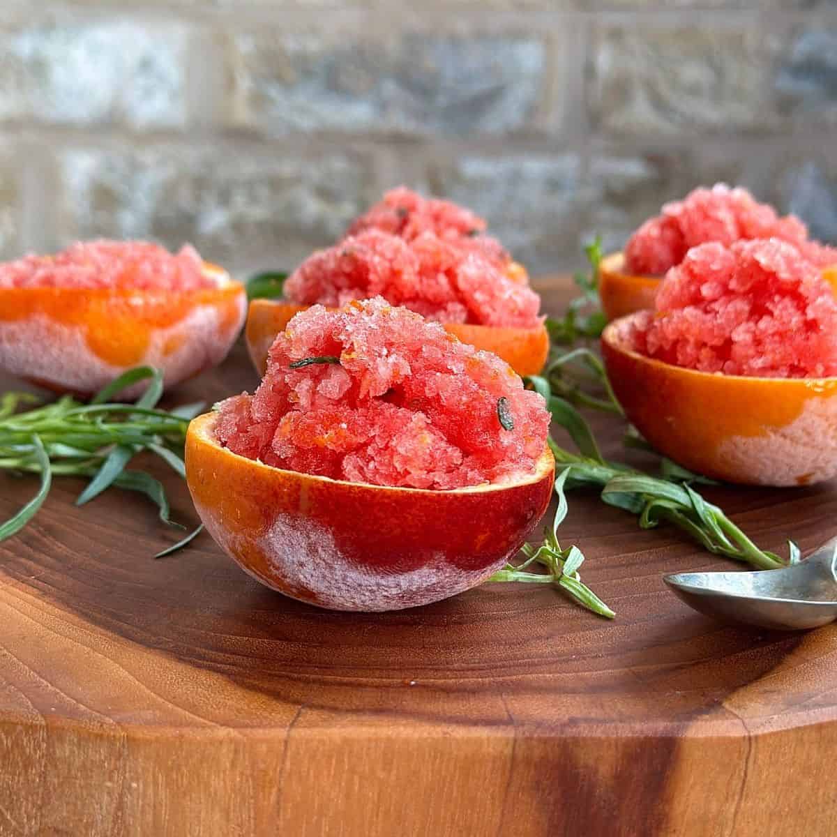 Blood orange granita served in orange shells with fresh tarragon on wooden board.
