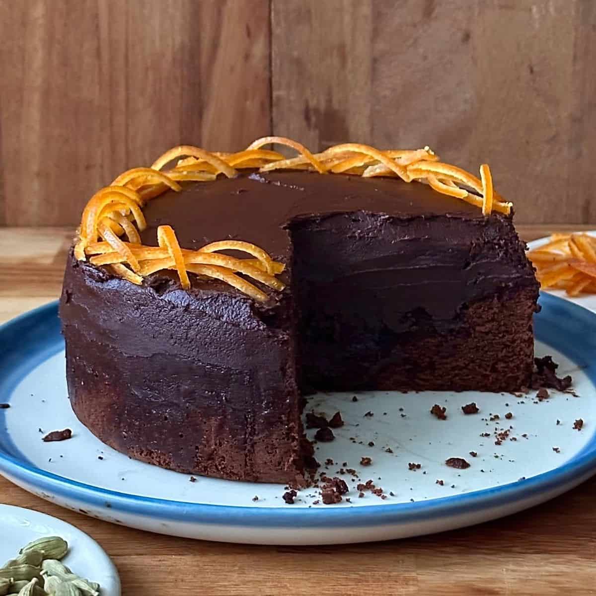 A dark chocolate mud cake garnished with thin strips of candied orange sitting on a white plate with a blue edge. A slice has been removed from the cake.