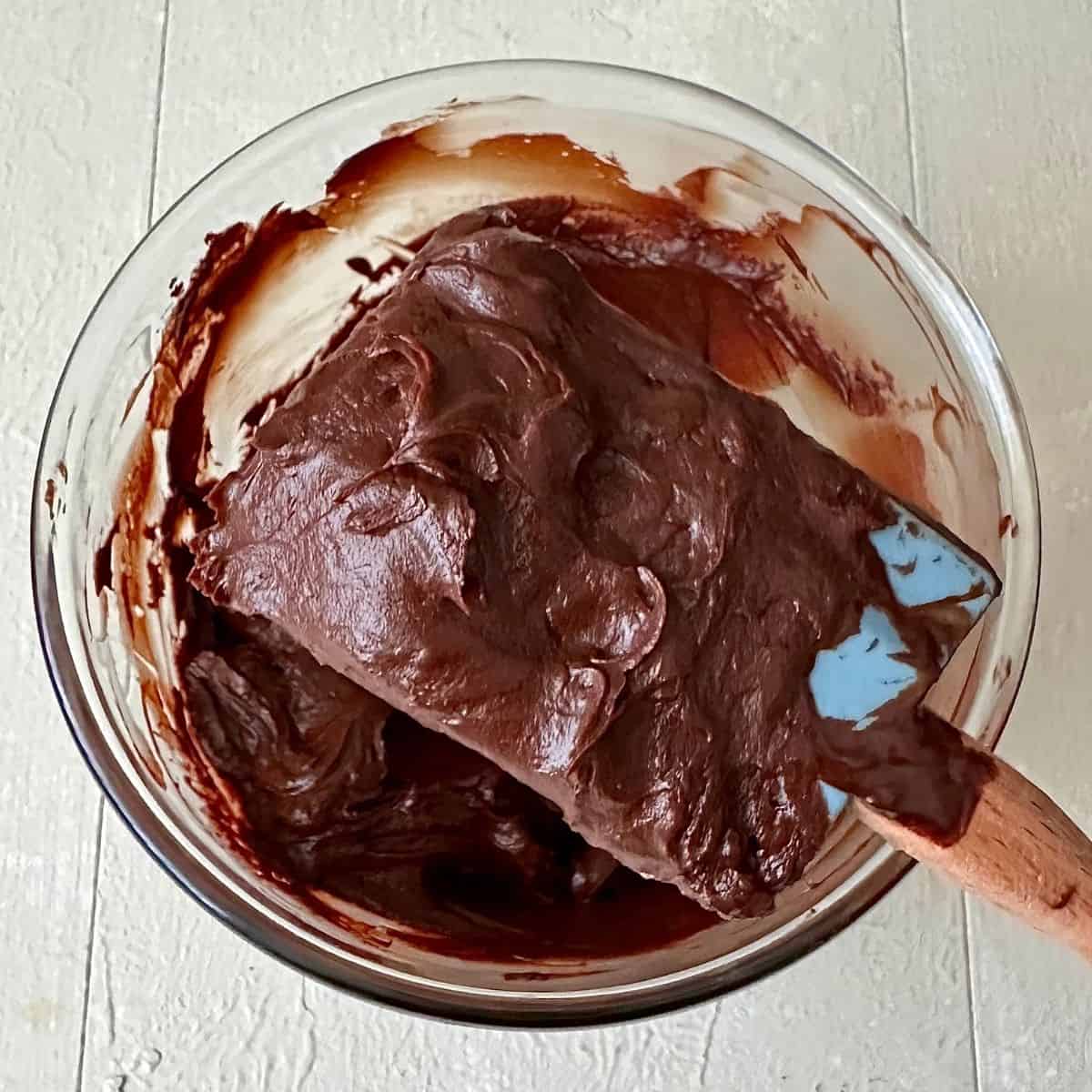Dark chocolate ganache on a spatula held over a glass bowl containing the ganache for a dark chocolate mud cake.