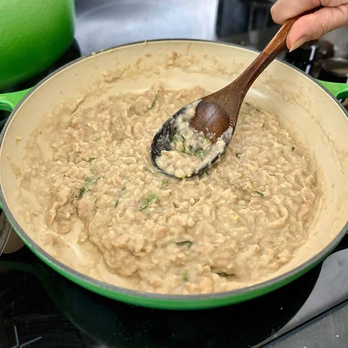 A large, shallow green pot containing mushy haricot beans. A back of a wooden ladle is being used to break down haricot beans to the mushy state.