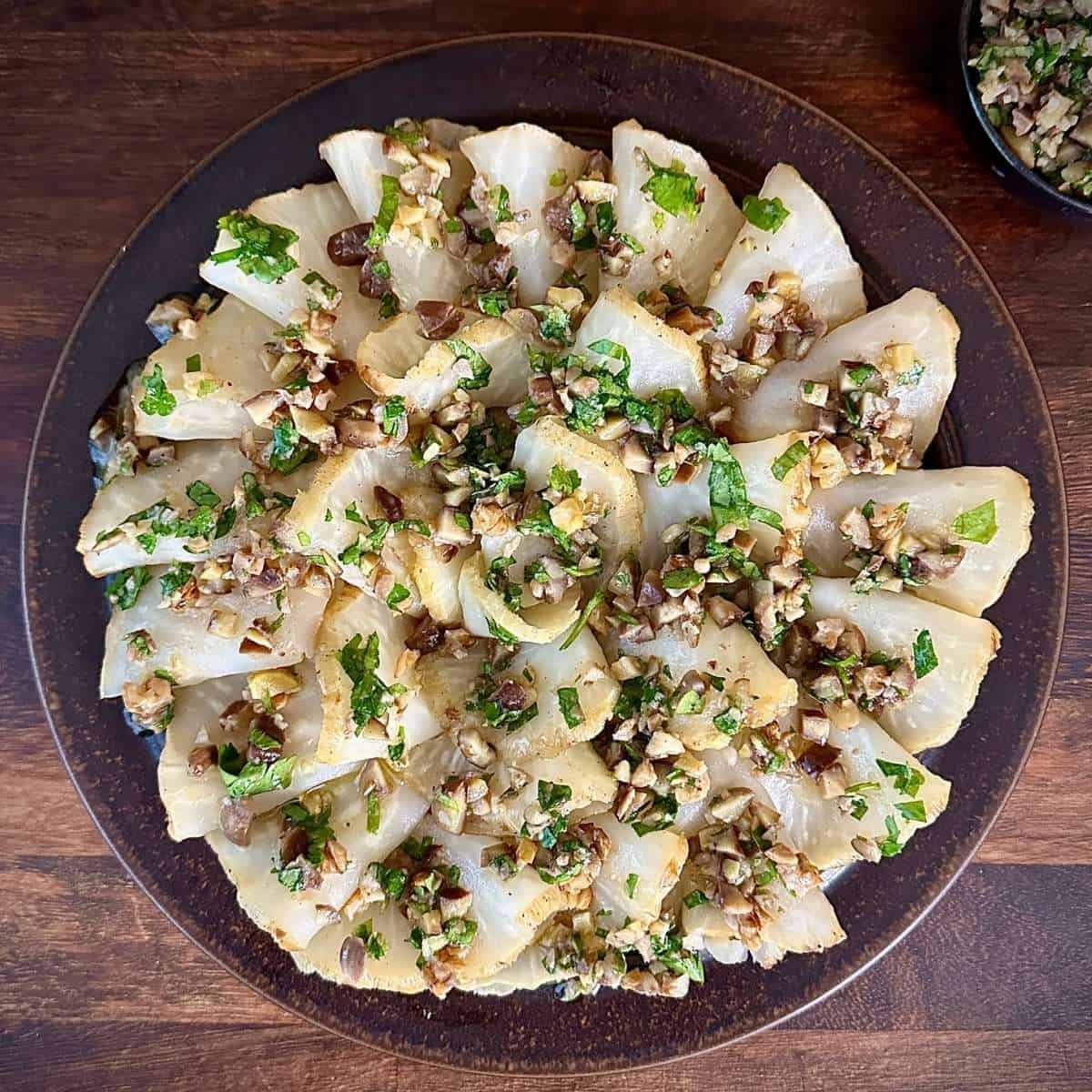 Thin Roasted celeriac slices on as rustic brown plate with an orange, walnut and chestnut and parsley dressing drizzled over.