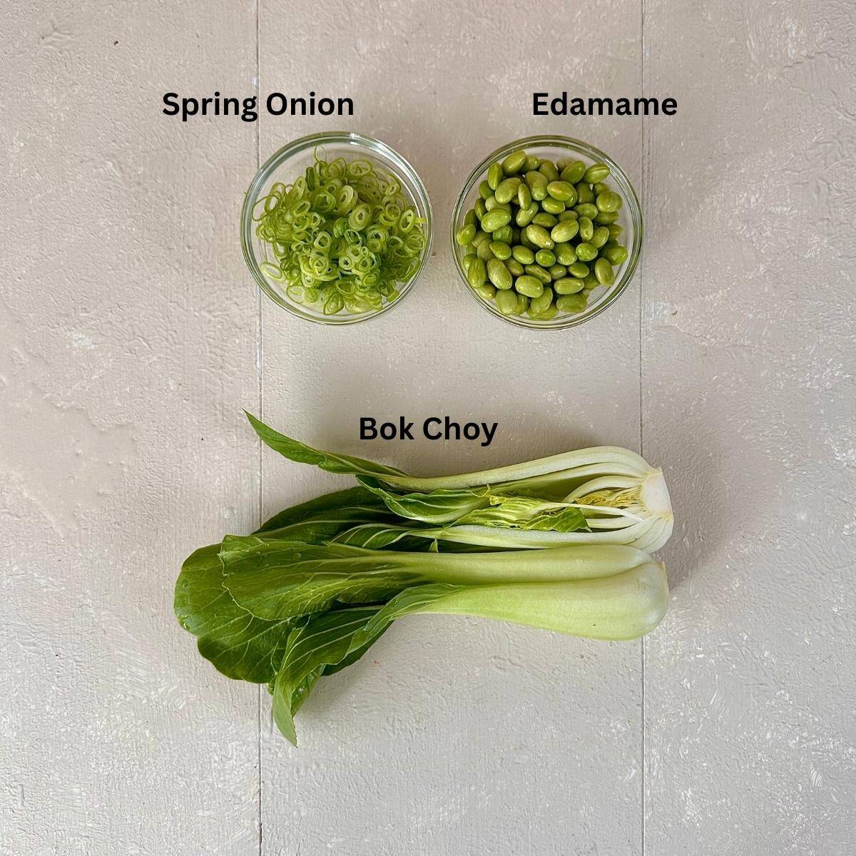Ingredients for veggie ramen garnish presented on a white background. Bok choy and sliced spring onions and edamame in small glass dishes. The ingredients are annotated.