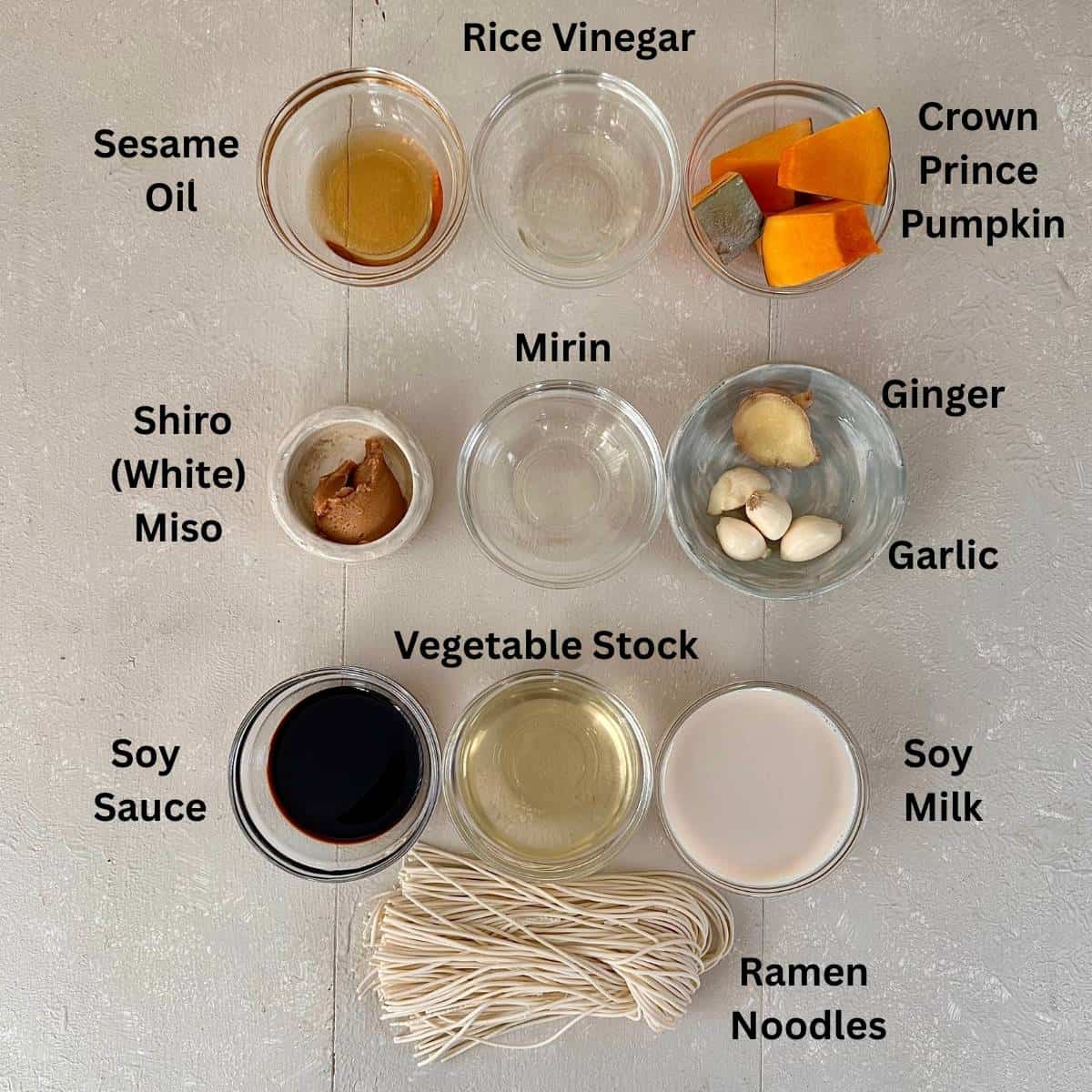 Ingredients for veggie ramen with miso and pumpkin broth laid out on a white surface in small glass dishes, including pumpkin, miso, garlic, ginger, soy sauce, stock, noodles and soy milk. Ingredients are annotated.