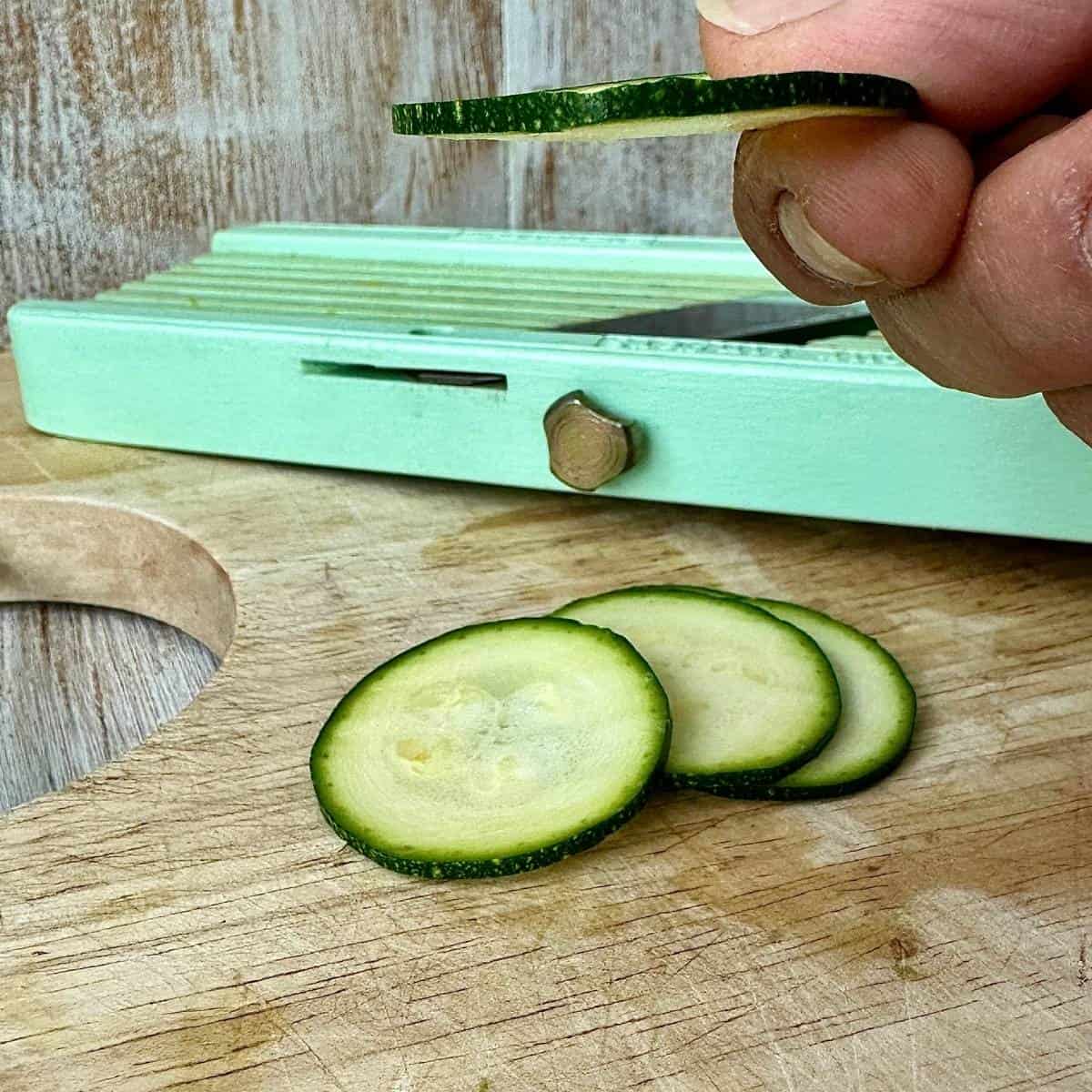 Slices of courgette lying on a wooden chopping board, having been sliced with a mandolin slicer. A hand holds one slice to show its thickness of 3mm.