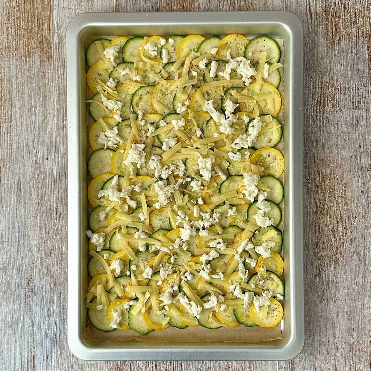 Thin courgette slices arranged in a baking tray overlapping each other to form a sheet. Sprinkled with grated mozzarella and cheddar cheese, ready to bake. For cottage cheese wrap with a courgette base.