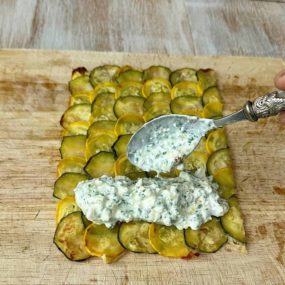 A courgette wrap sheet with cottage cheese and chives filling at one end, ready to roll into a wrap. A spoon is held over the wrap with cottage cheese and chives falling off it.