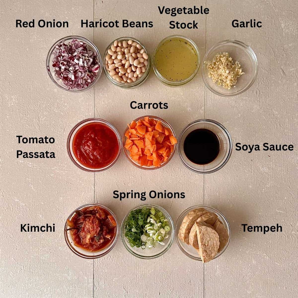 Prepared ingredients for kimchi white bean stew with tempeh crumble. Ingredients are in small glass dishes and are annotated.