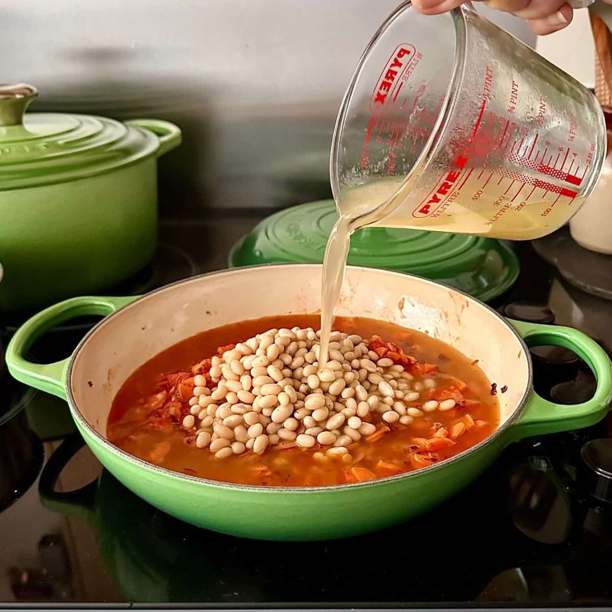 Pouring stock into kimchi white bean stew in a green casserole dish on the hob.