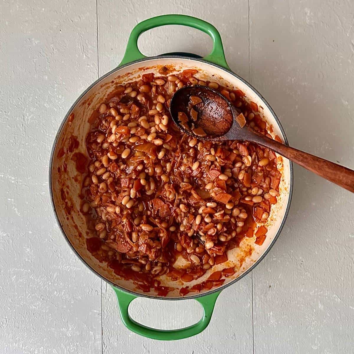 Kimchi white bean stew in a green casserole dish with a wooden spoon, viewed from above.