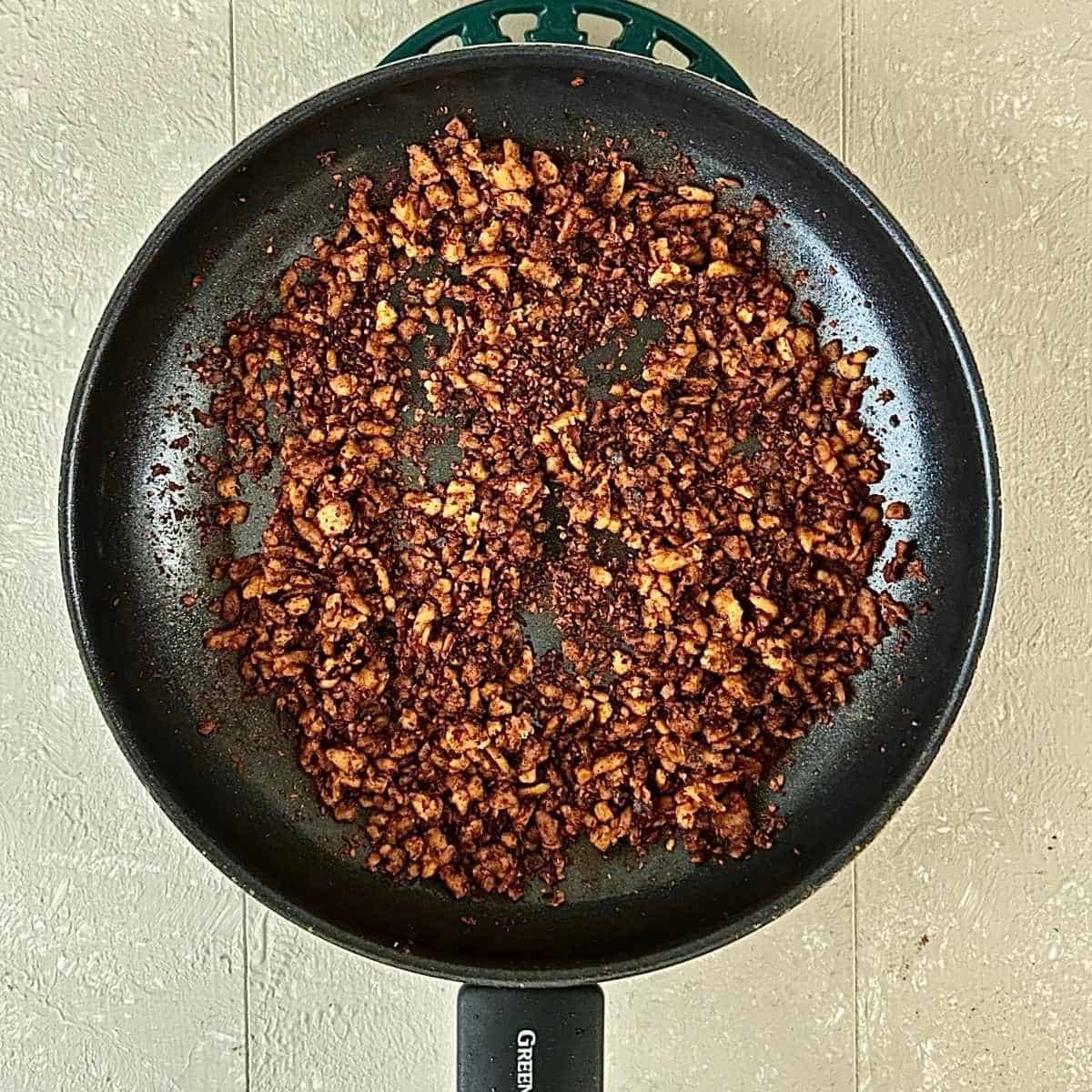 A large frying pan containing fried grated tempeh marinated in soya sauce and kimchi juice. Golden brown in colour.