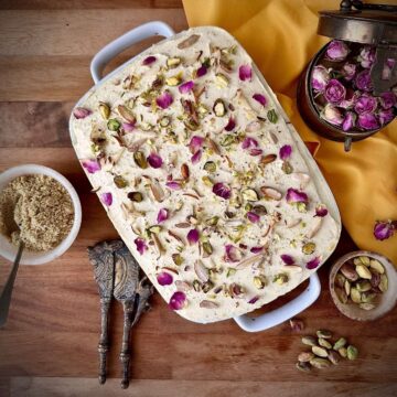 Overhead view of no-bake thandai cream cake in a white dish, garnished with pistachios, almonds and dried rose petals.