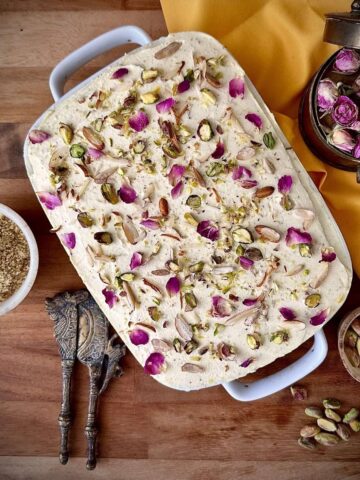 Overhead view of no-bake thandai cream cake in a white dish, garnished with pistachios, almonds and dried rose petals.