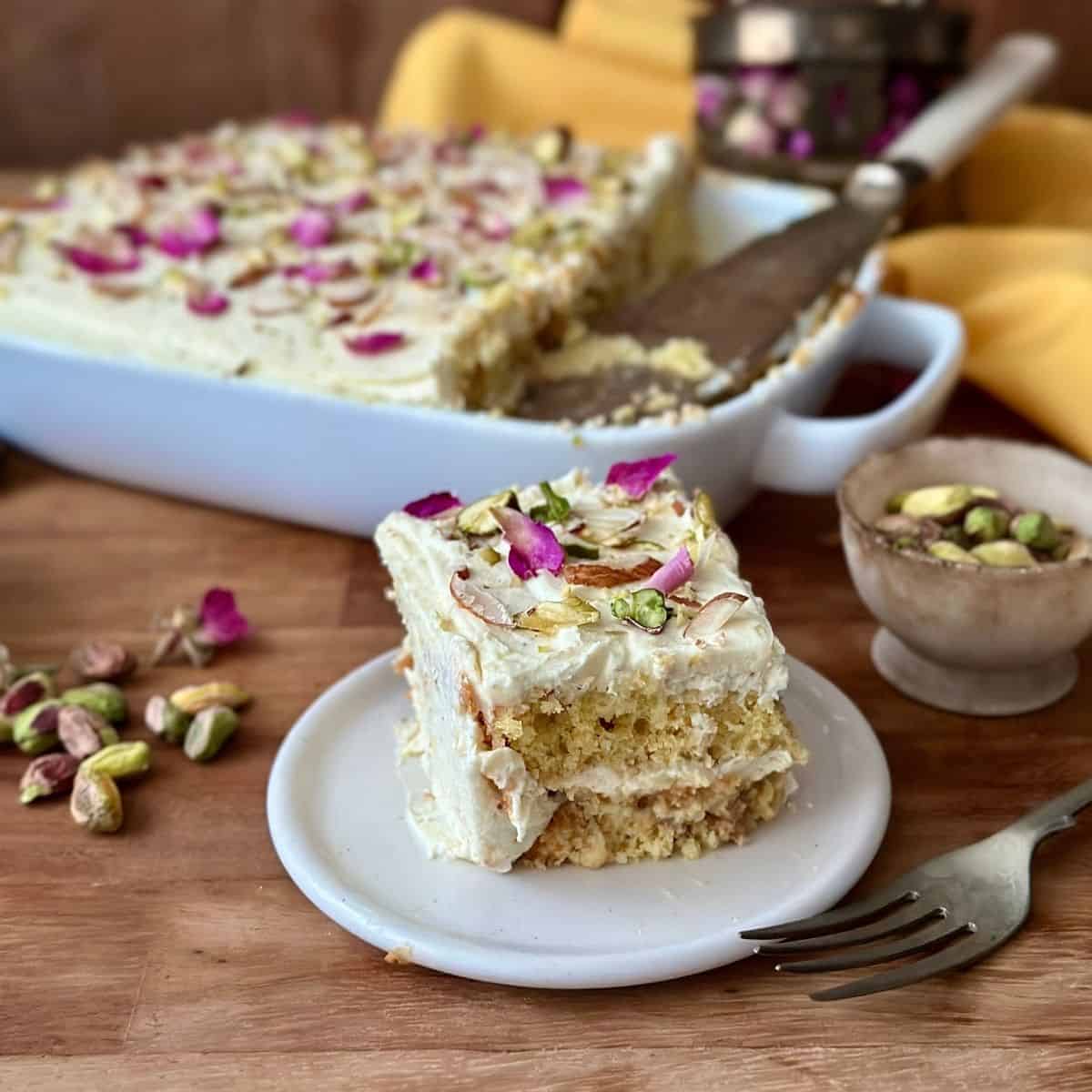 Slice of no-bake thandai cream cake topped with pistachios and rose petals, showing soft rusk layers and spiced mascarpone filling.