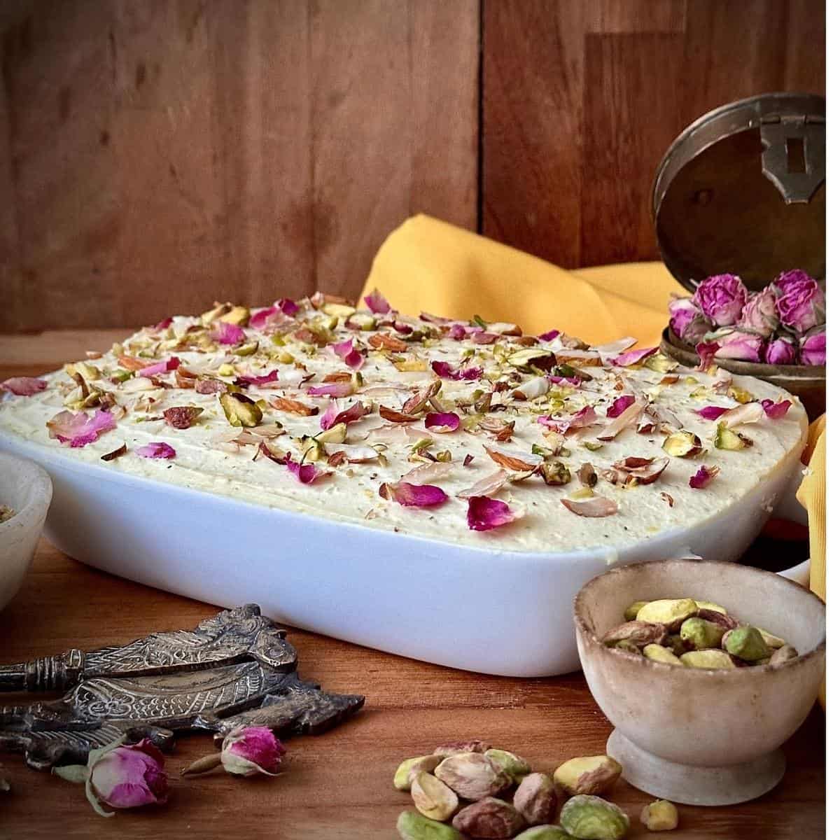 A no-bake thundai cream cake in a shall rectangular dish, garnished with pistachio and almond pieces and dried rose petals