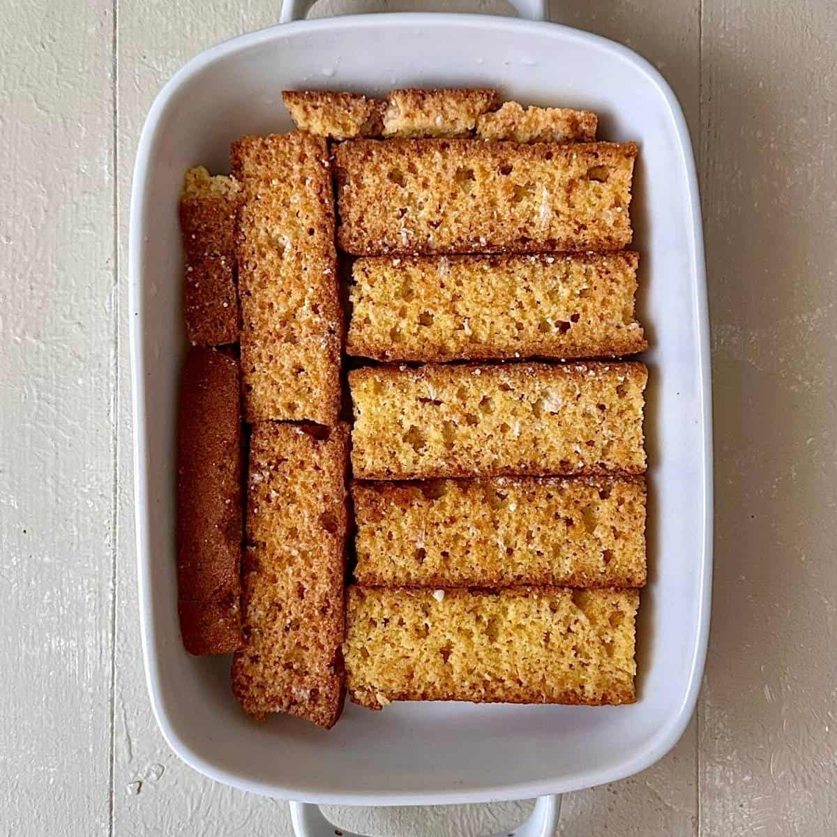 A shallow rectangular, white serving dish containing a layer of cake rusk soaked in milk. For a no-bake thundai cream cake.