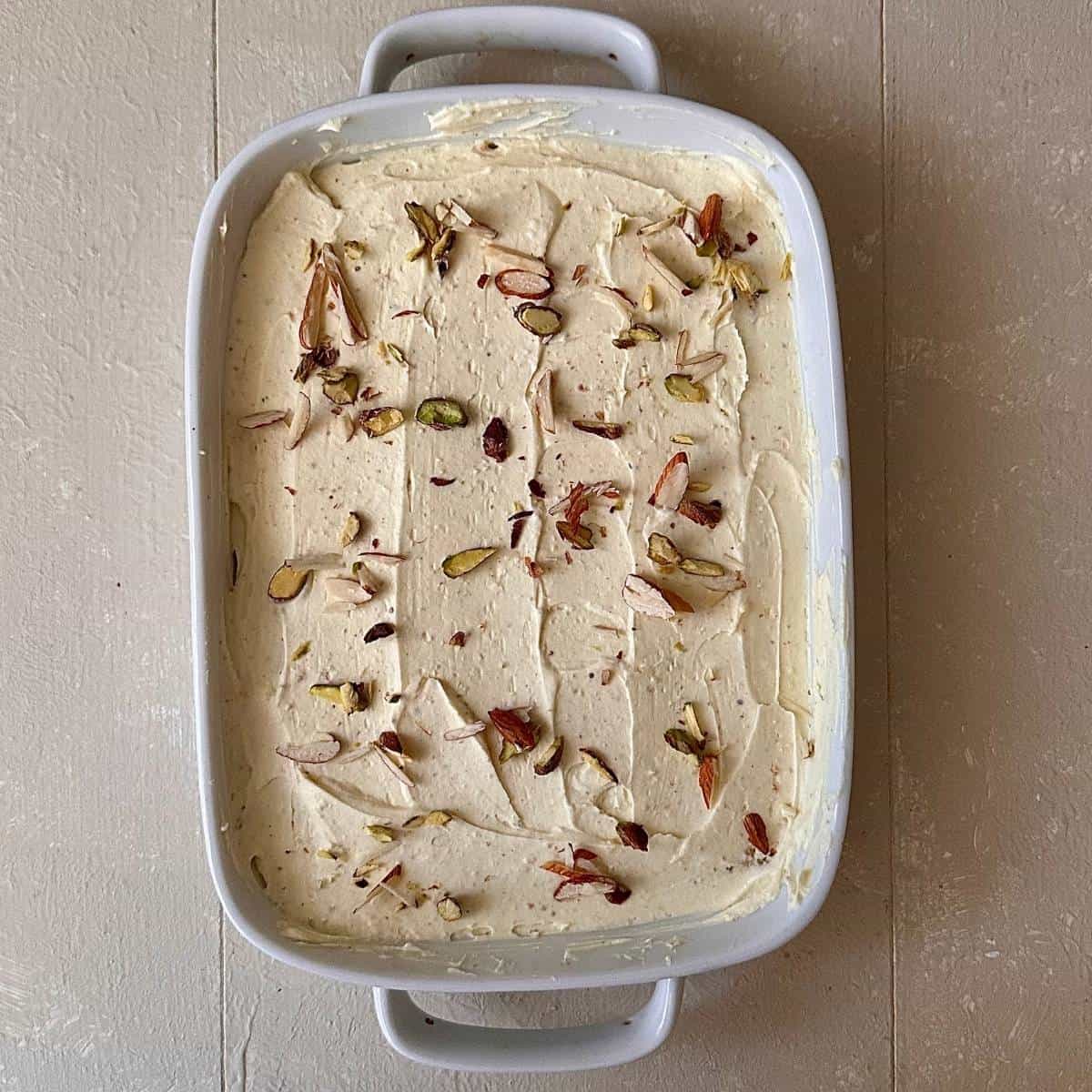 A shallow white rectangular dish with a layer of cake rusk covered in a layer of thundai cream for thundai cream cake.