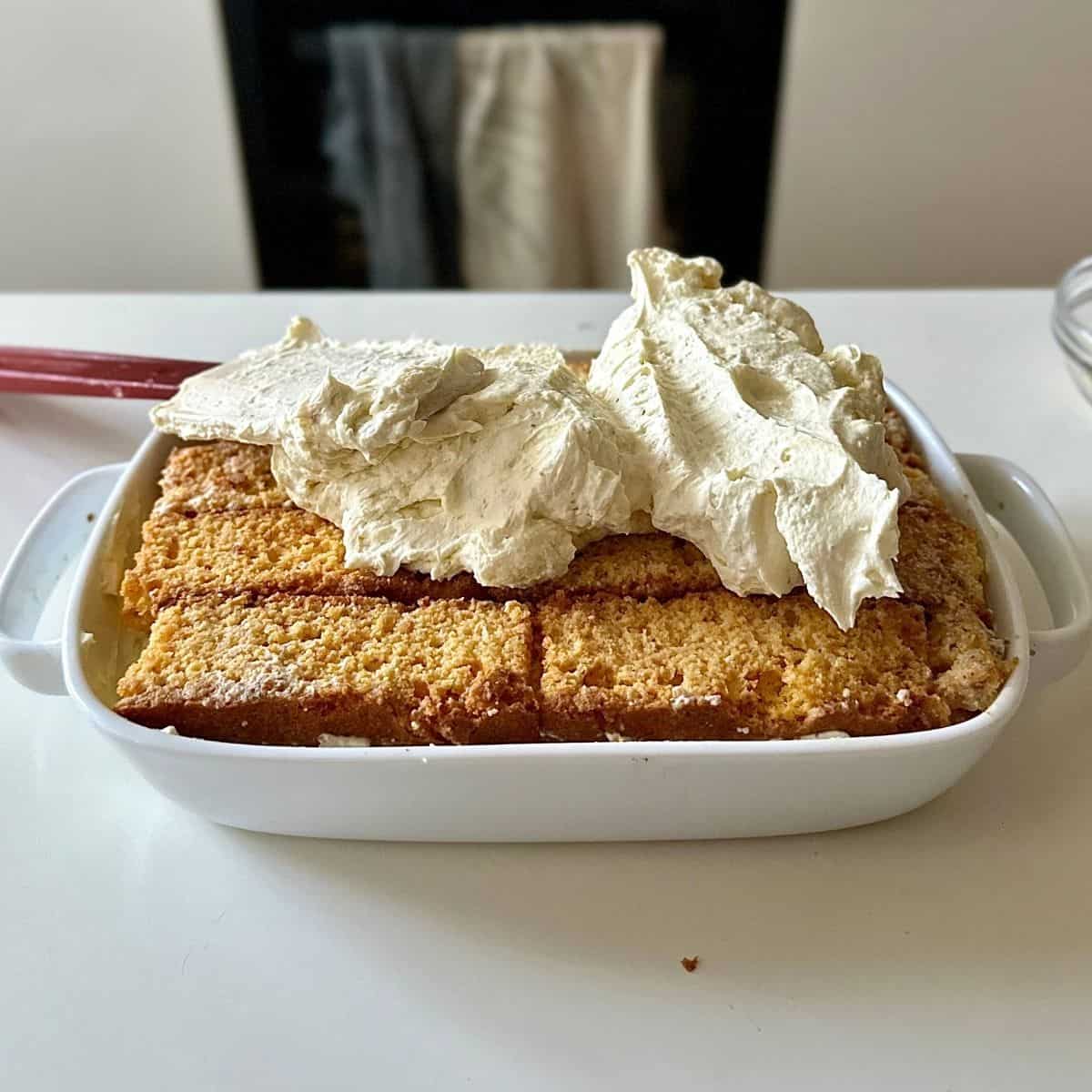 A shallow rectangular white serving dish containing cake rusk and thundai cake cream layer. A top layer of cream sits on top of cake rusk, ready to smooth out flat. A spatular sits on the dish.