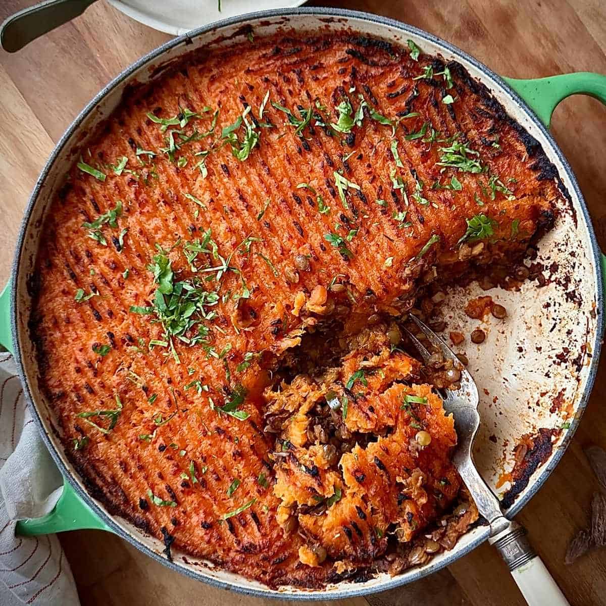A large shallow green casserole dish containing sweet potato lentil shepherd's pie garnished with coriander. A portion has been removed.