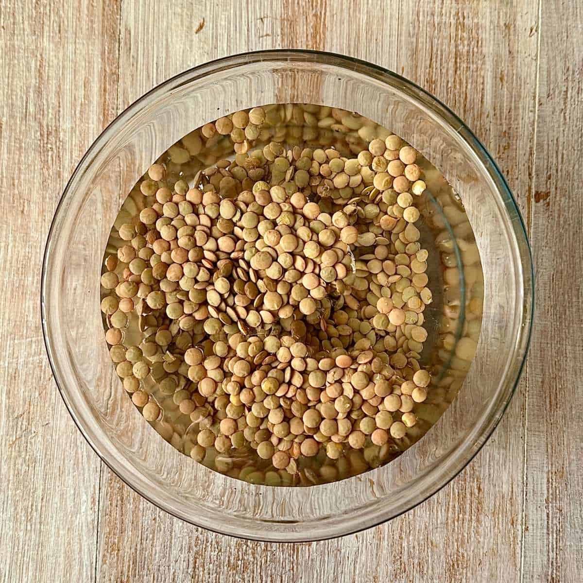 A glass bowl containing green lentils soaking in water.