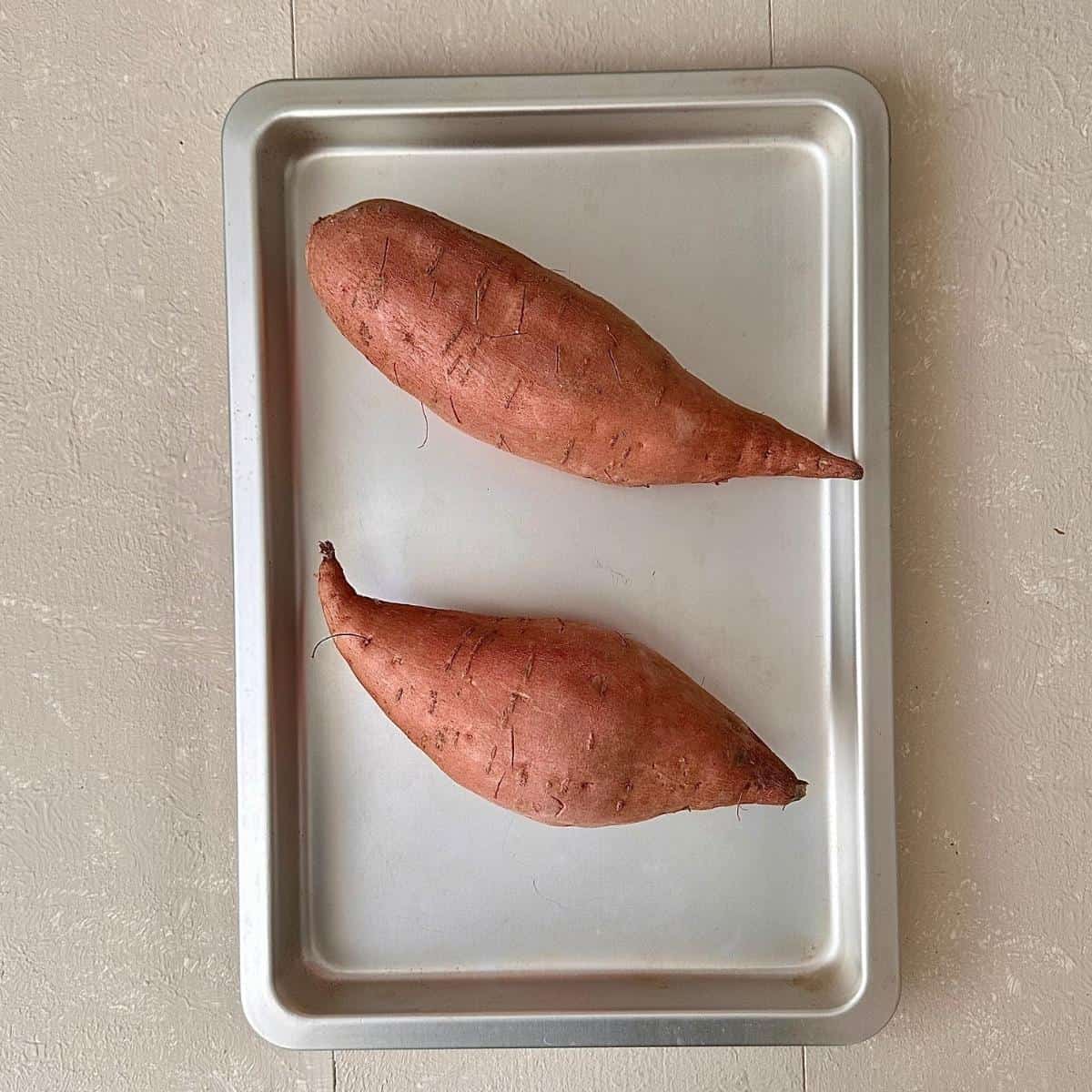 Two whole sweet potatoes in a baking tray, ready to bake in the oven.
