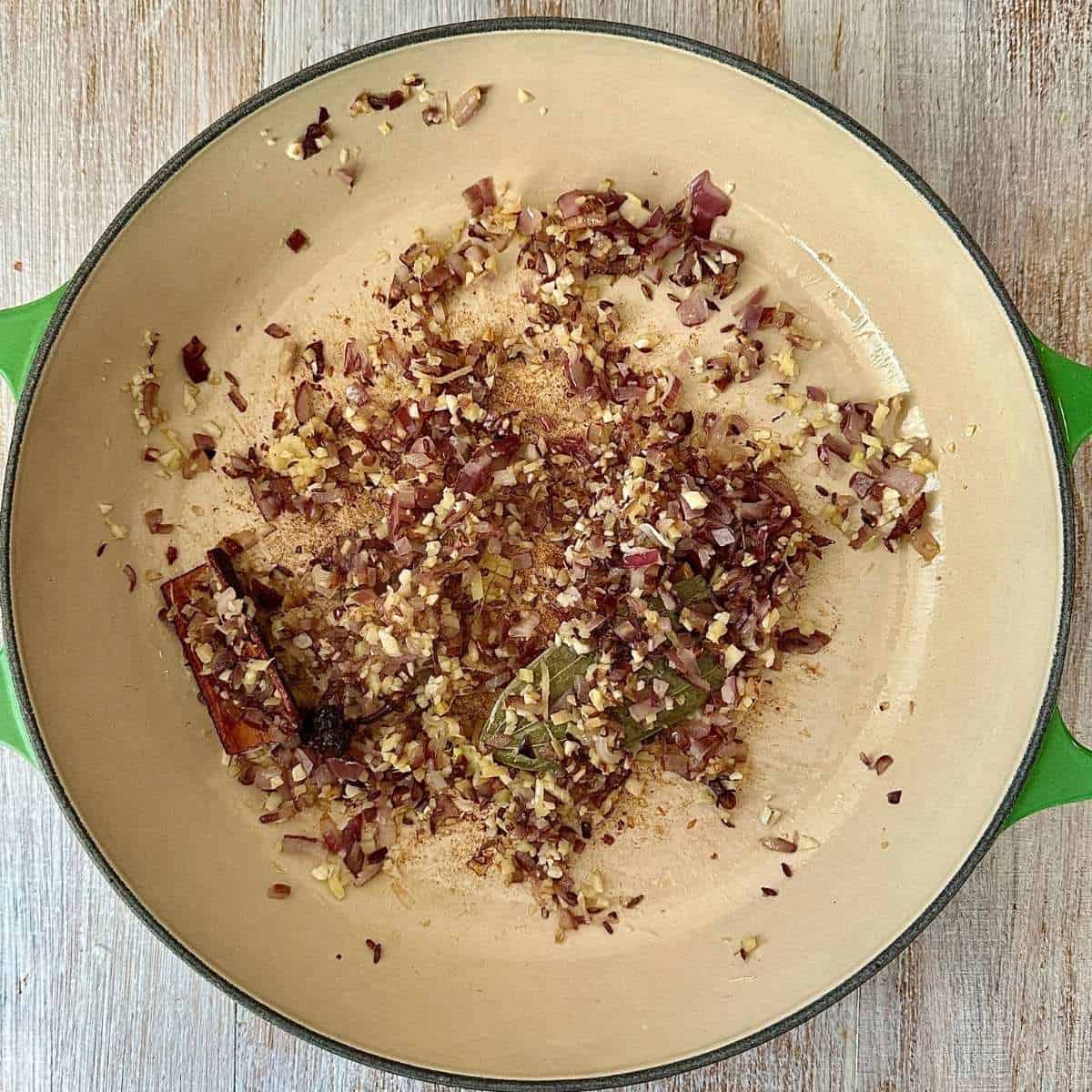 A large green casserole dish containing fried finely chopped onion, garlic and ginger, tej patta, cassia bark and cloves. For sweet potato lentil shepherds pie.