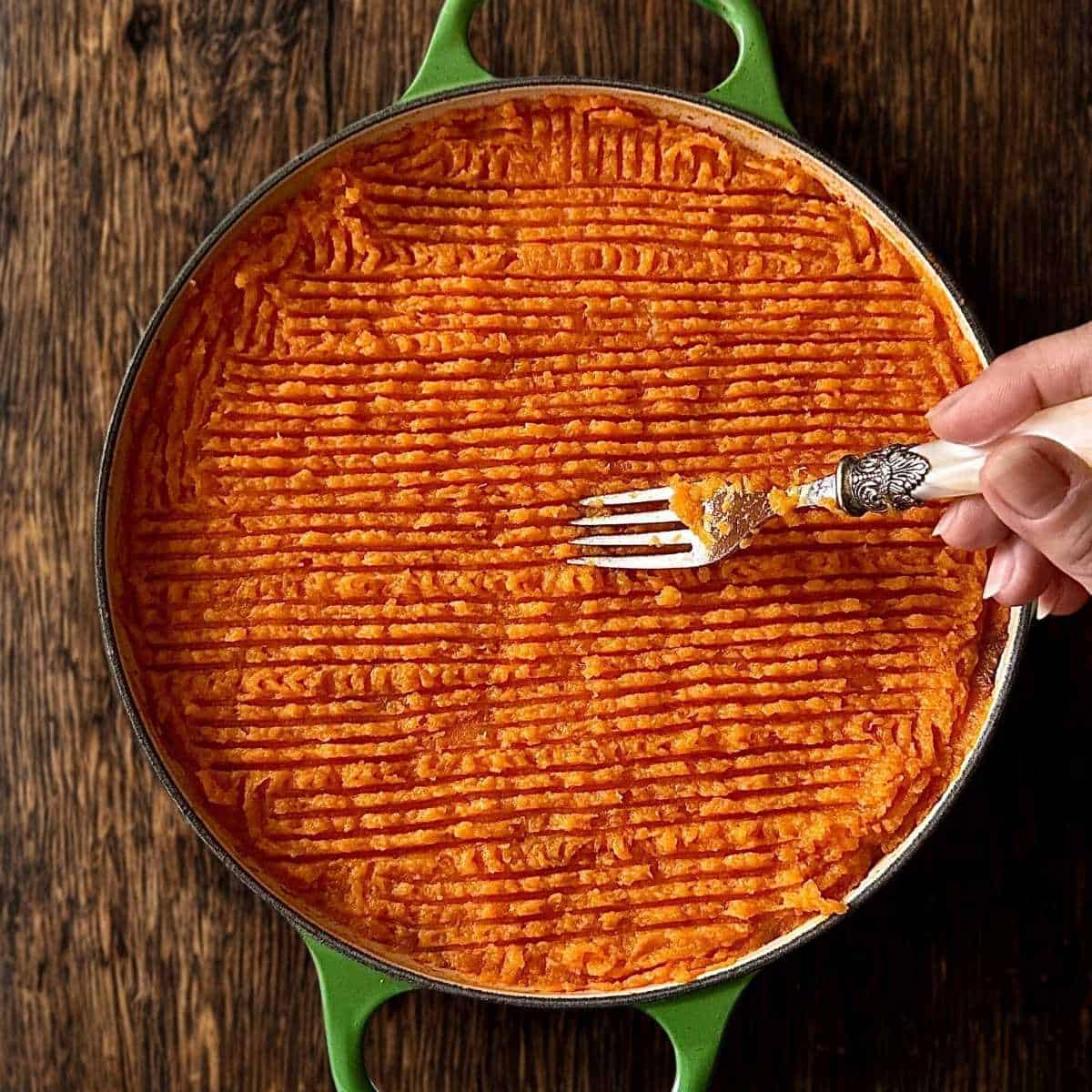 A sweet potato lentil shepherd's pie in a shallow green casserole dish. A Fork is being drawn across the surface of the mashed sweet potato to form small ridges.