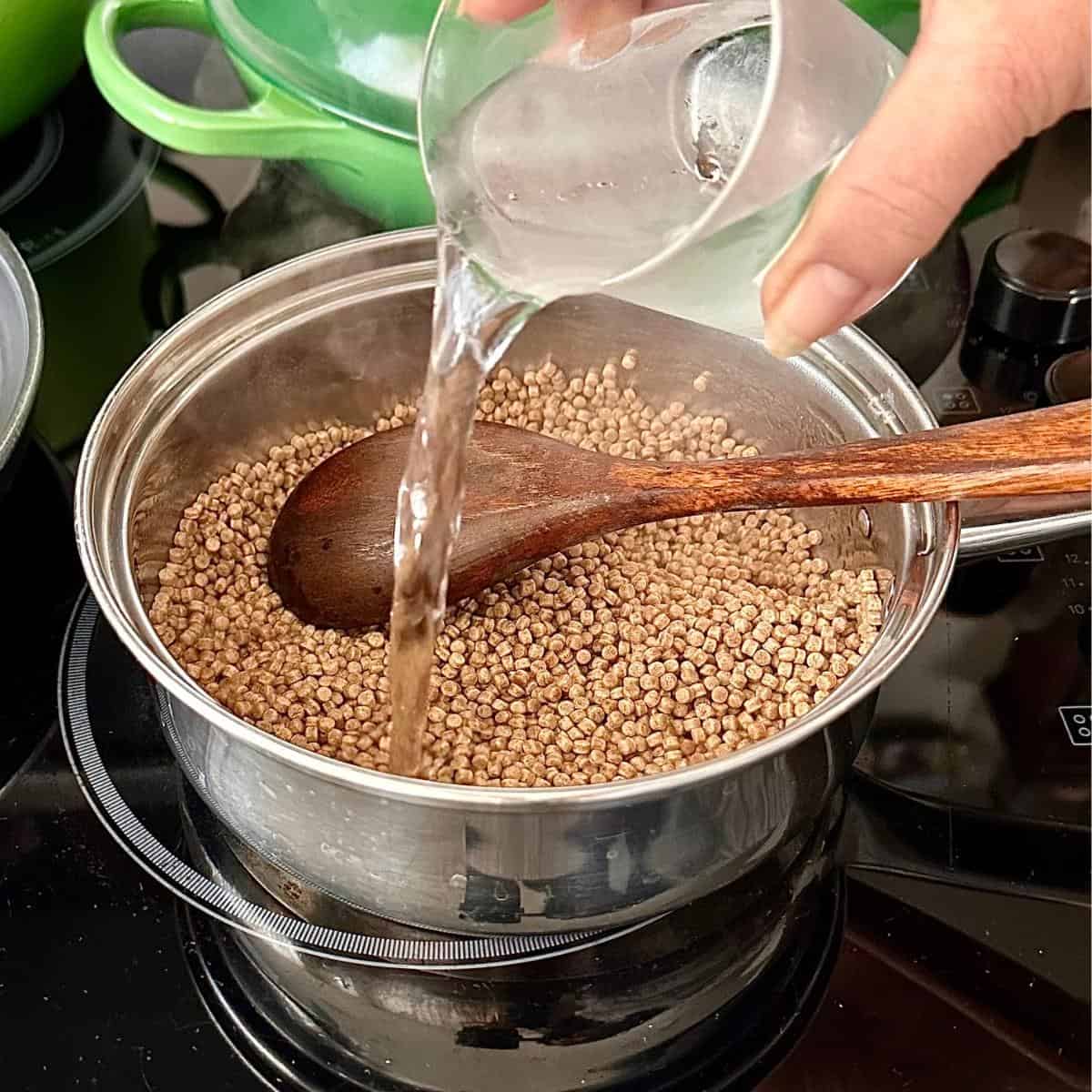 A saucepan containing giant wholewheat couscous for za'atar halloumi couscous salad. A hand is pouring water from a beaker into the pot.