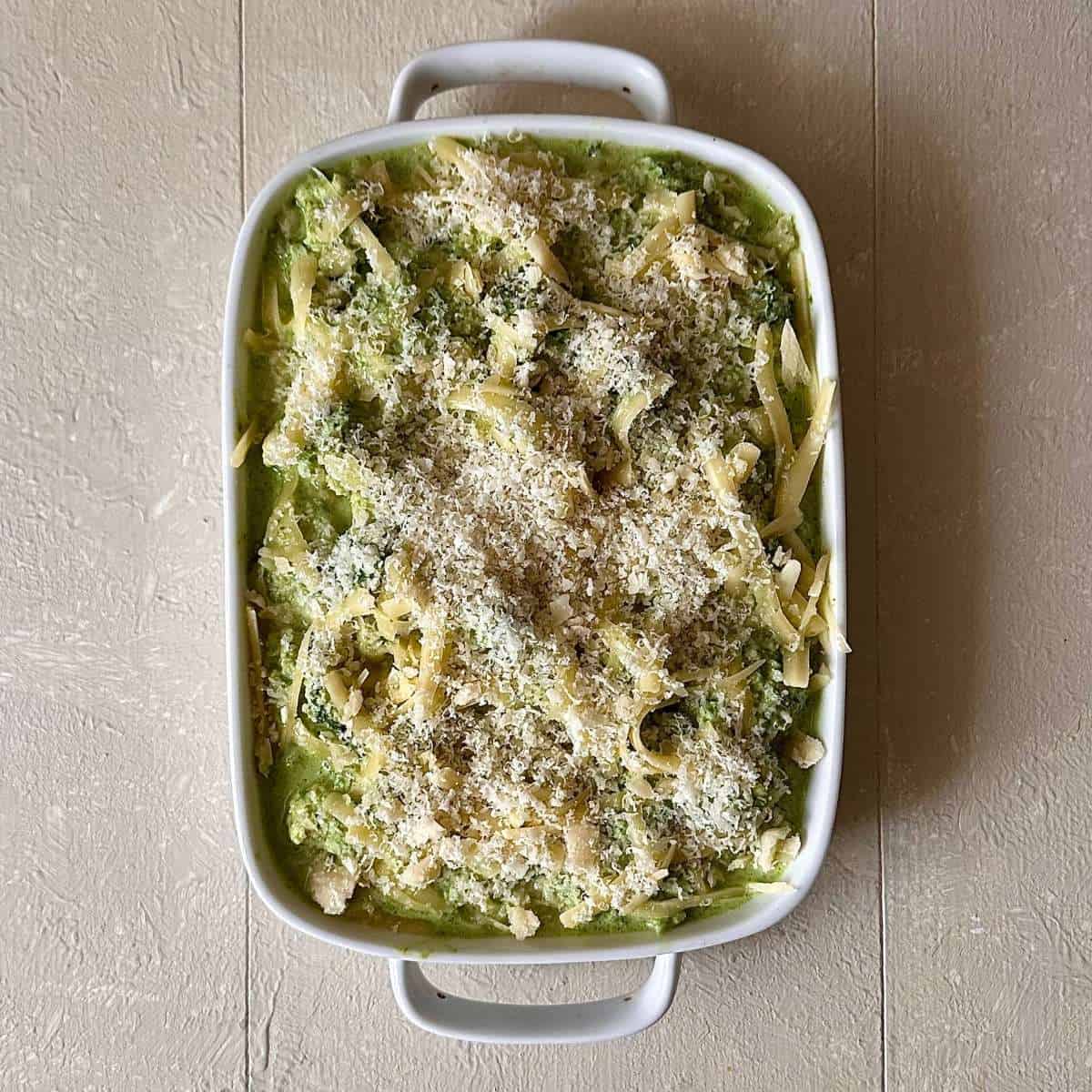 An oven-proof dish containing wild garlic cauliflower cheese with grated cheddar cheese and parmesan cheese, ready for bake in the oven.