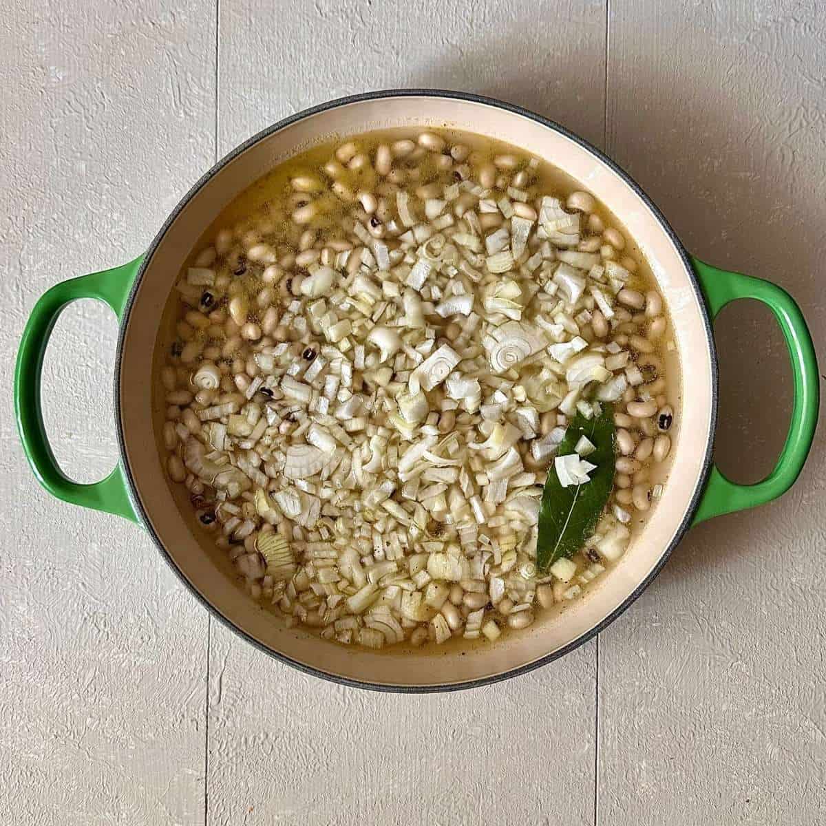 A large green casserole containing roughly chopped onion, black eyed peas, bay leaf and vegetable stock for black eyed peas with smoked tofu recipe.