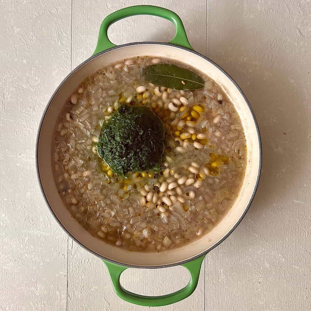 A large green casserole containing roughly chopped onion, black eyed peas, bay leaf and vegetable stock. Pureed wild garlic sits on top ready to incorporated.