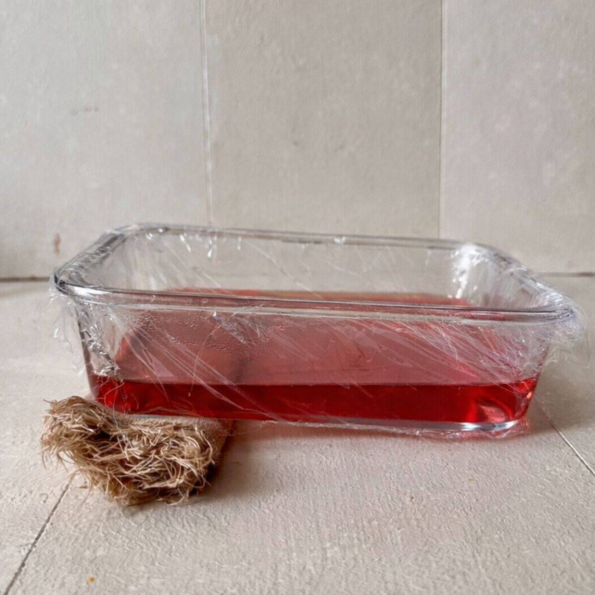 A cling film-lined glass dish containing flowering current jelly mix. The bowl has a small rolled towel under one end to enable the jelly to set at an angle.