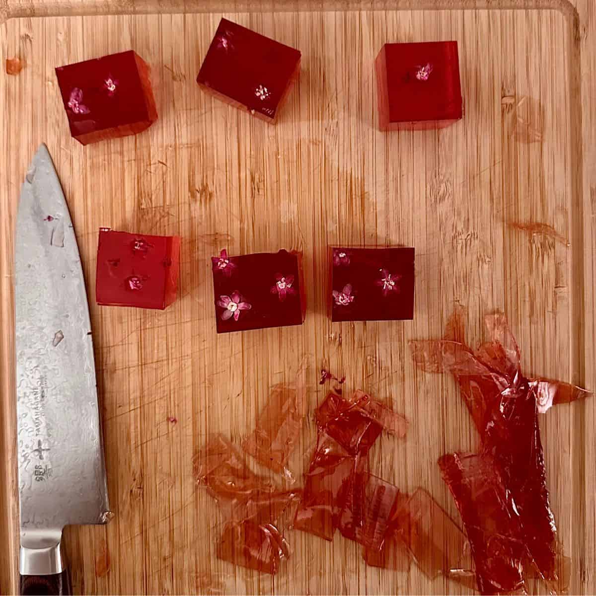 Six flowering current jelly cubes, about 1 inch in size sitting on a wooden chopping board. A knife and off-cuts of jelly sits next the cubes.