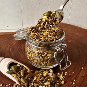 Super seed mix in a glass jar with a spoonful lifted above, showing toasted pumpkin seeds, sunflower seeds, sesame, linseeds and spices.