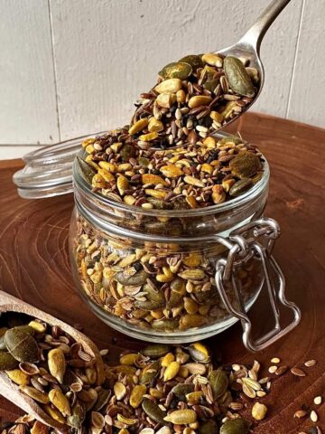 Super seed mix in a glass jar with a spoonful lifted above, showing toasted pumpkin seeds, sunflower seeds, sesame, linseeds and spices.