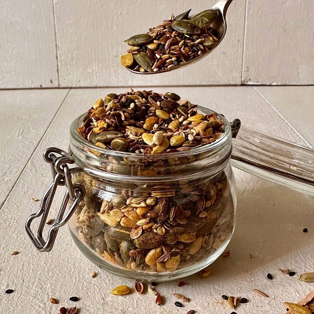A glass jar containing supper seed mix. A teaspoon with seed mix is held above the jar.