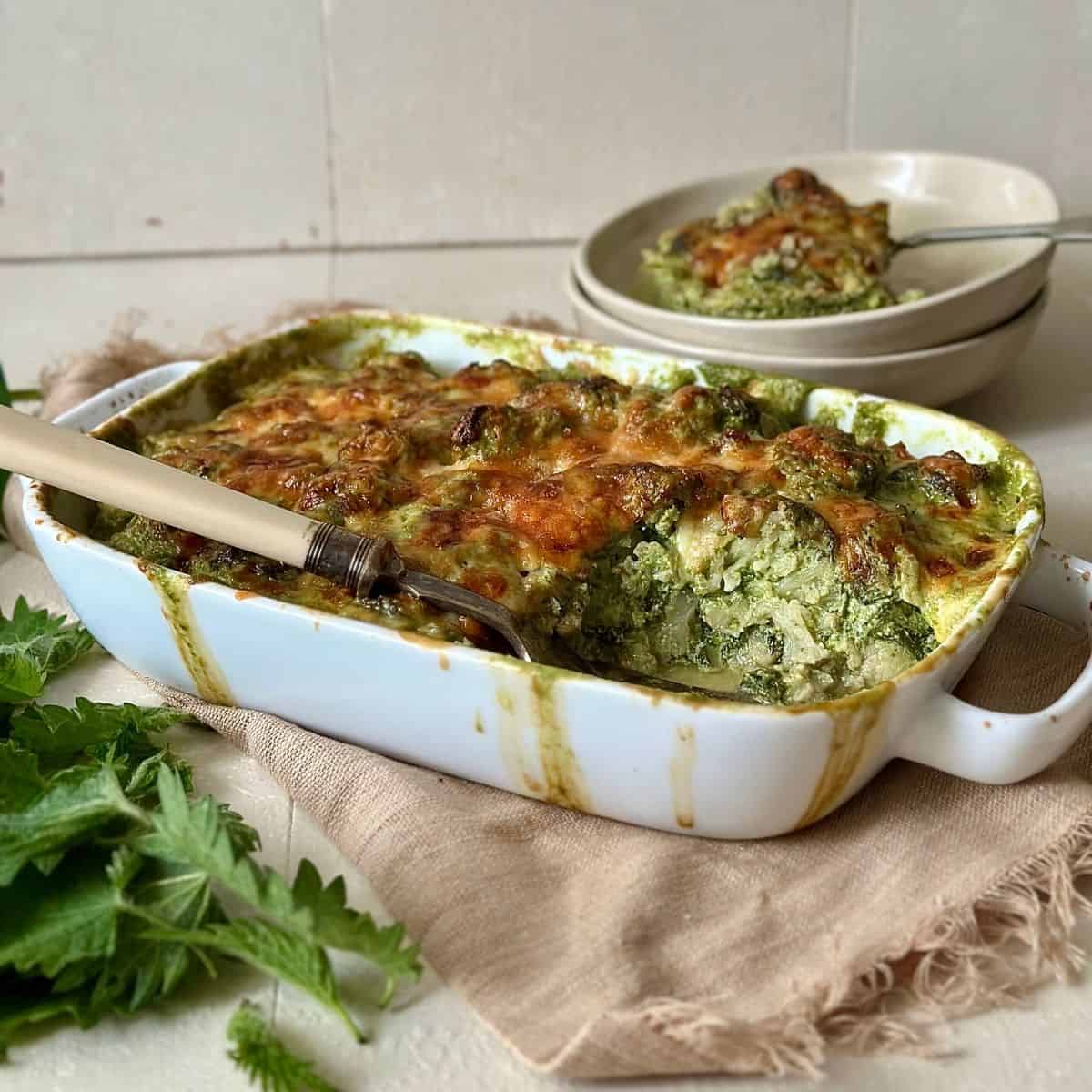 Wild garlic cauliflower cheese in a white oven-proof dish. A portion has been removed and sits on a plate in the background.
