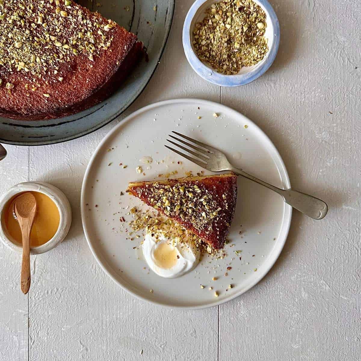 A slice of orange polenta cake scattered with chopped pistachio and served with creme fraiche and honey. On a small plate with a Fork.
