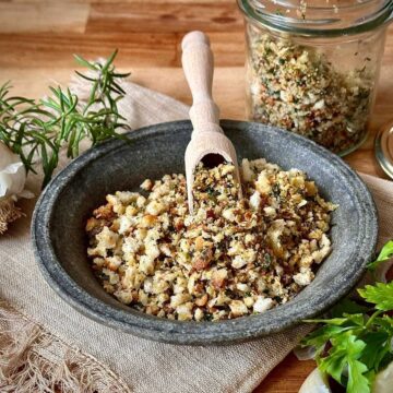 A stoneware bowl containing pangrattato crispy Italian breadcrumbs. A small wooden scoop rests on the pangrattato.