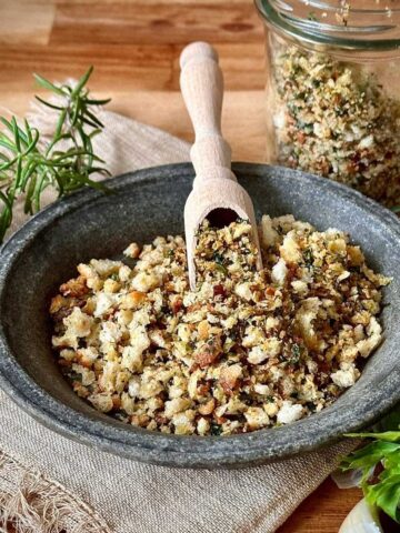 A stoneware bowl containing pangrattato crispy Italian breadcrumbs. A small wooden scoop rests on the pangrattato.