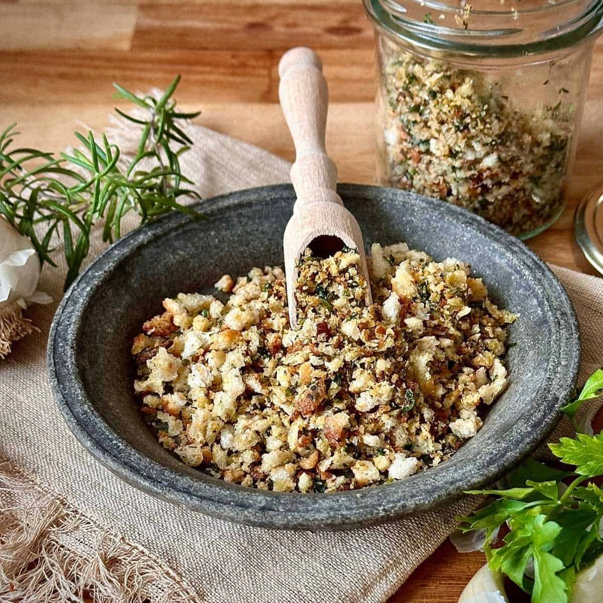 A stoneware bowl containing pangrattato crispy Italian breadcrumbs. A small wooden scoop rests on the pangrattato.
