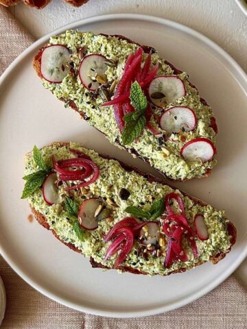 A shallow beige plate with two pieces of sourdough toast spread with smashed edamame and kale stems, garnished with pickled red onion, radish and mint.