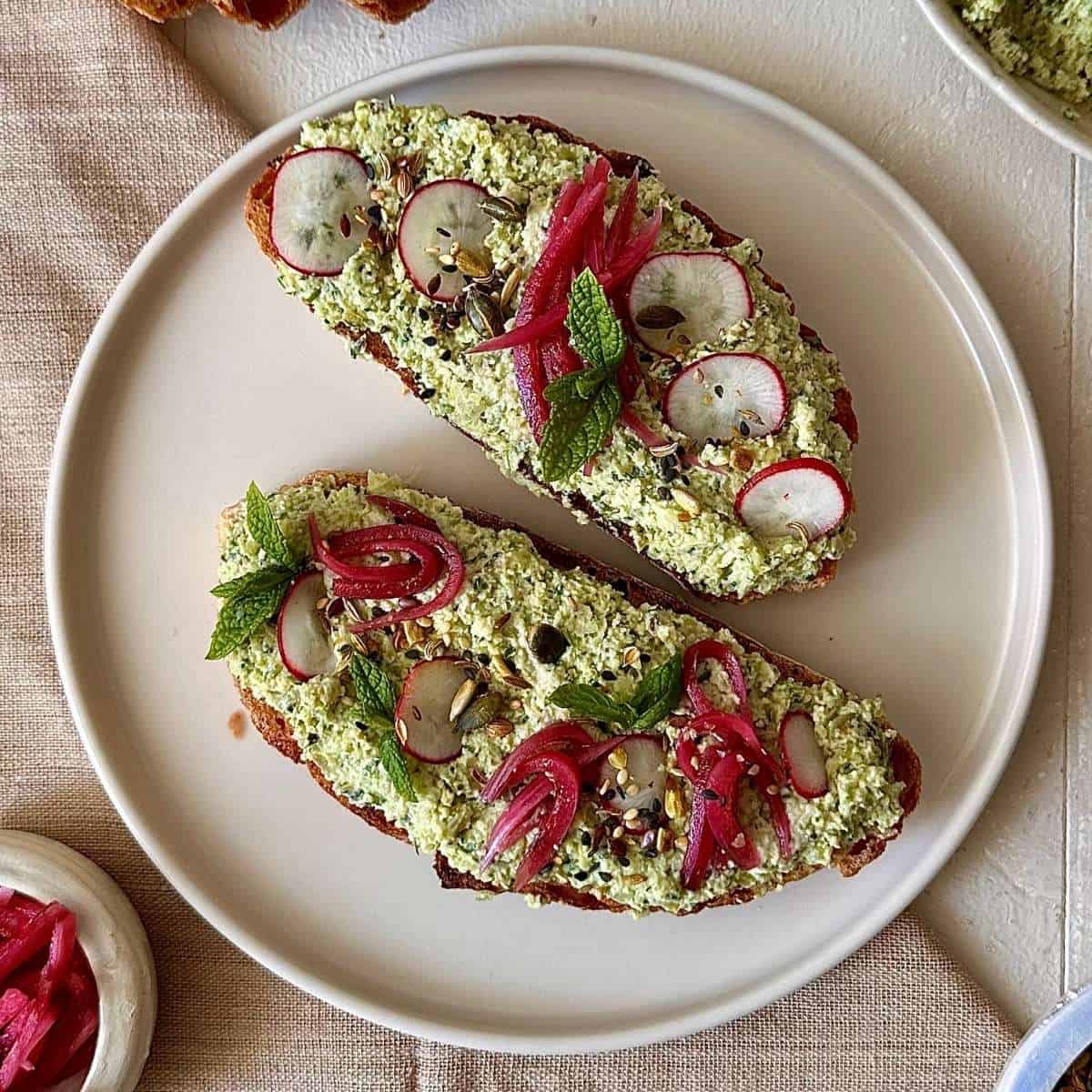 A shallow beige plate with two pieces of sourdough toast spread with smashed edamame and kale stems, garnished with pickled red onion, radish and mint.