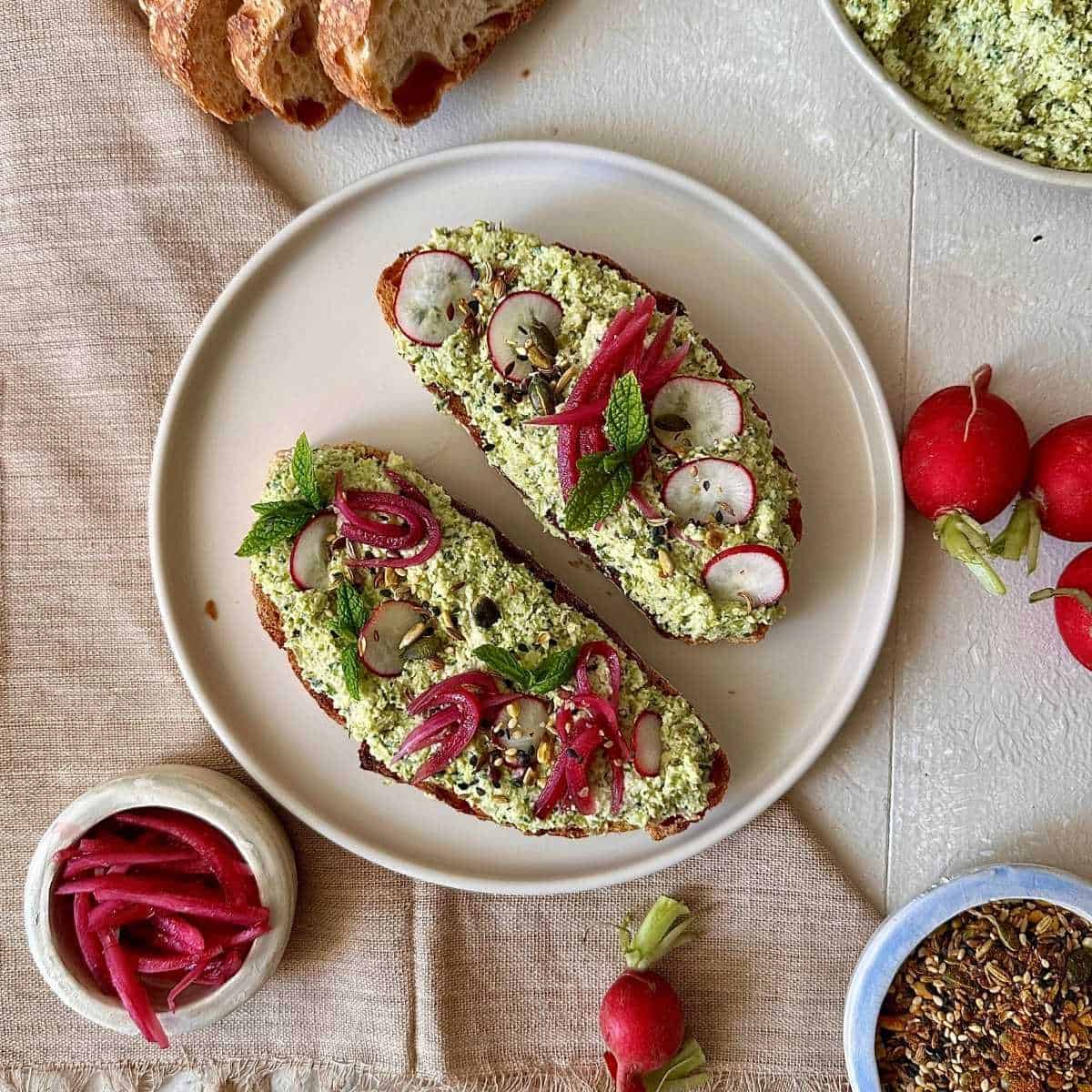 A beige plate with two slices of sourdough toast with smashed edamame and kale stems, garnished with pickled red onion, radish and mint.