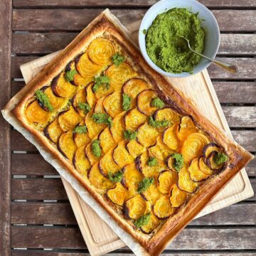 A beetroot tart garnished with beetroot leaf pesto, cut into 8 slices, sitting on parchment paper and a wooden chopping board. A bowl of beetroot leaf pesto sits next to the tart.